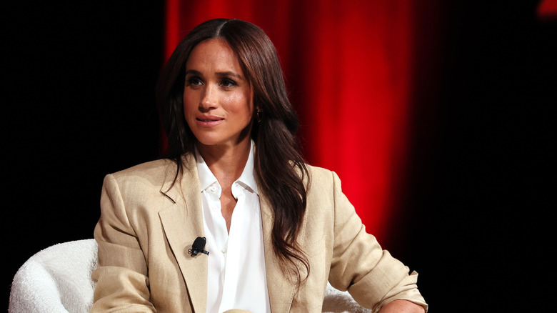 Meghan Markle listening onstage in a beige suit jacket and a white blouse