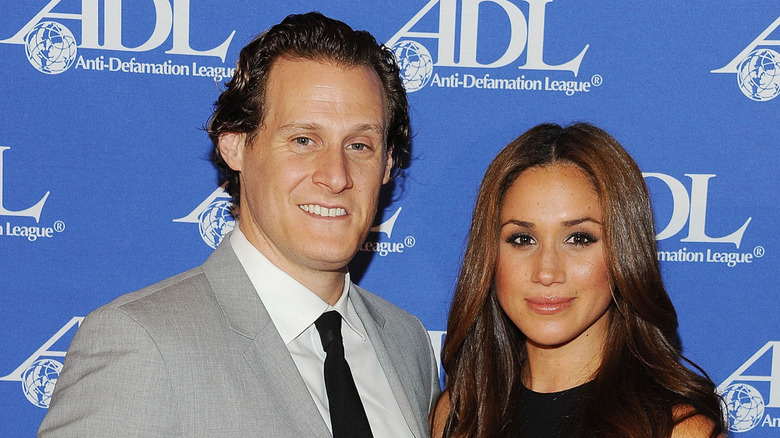 Trevor Engelson smiling and Meghan Markle smirking