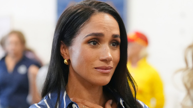Meghan Markle touches her neck while wearing small gold hoop earrings and a navy blue and white striped shirt.