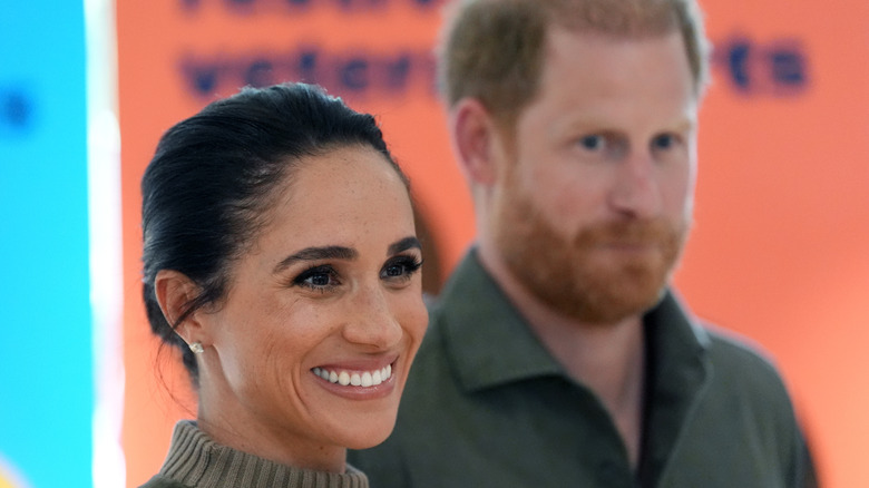 Meghan Markle wearing a green turtleneck and smiling with Prince Harry in the background.