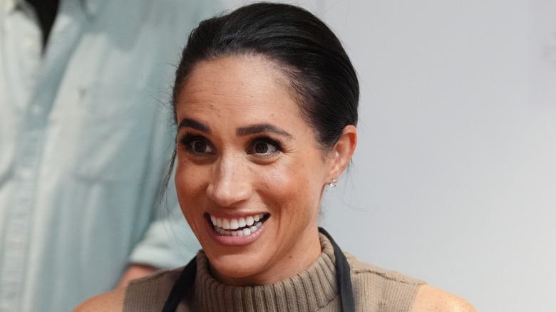 Meghan Markle in khaki turtleneck sleeveless shirt smiling.