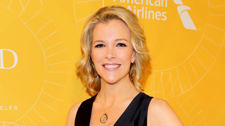 Megyn Kelly seated at her desk in a white dress looking away from the camera
