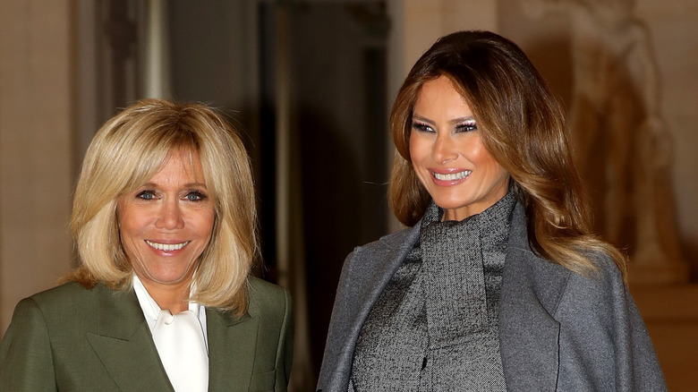 Brigitte Macron smiling in a green blazer and white collared shirt while standing next to a smiling Melania Trump who's wearing a silver shirt and coat at the Head of State partner's luncheon.