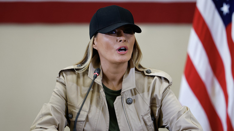 Melania Trump mouth open in a black baseball cap and tan jacket in front of an American flag