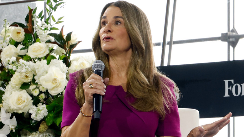 Melinda French Gates speaking into microphone in a purple dress next to some flowers