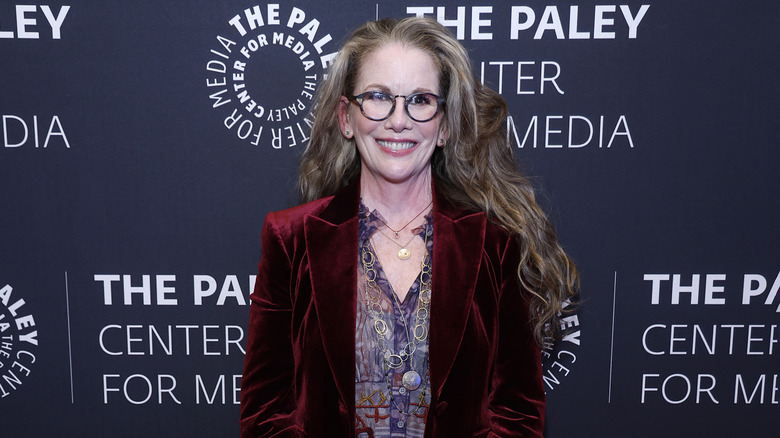 Melissa Gilbert smiling while posing in a maroon velvet blazer, patterned blouse, and glasses