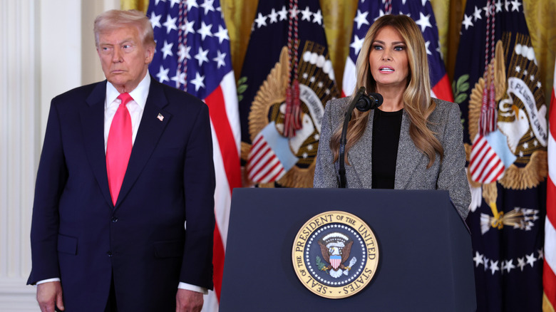 Donald Trump stands next to Melania Trump as she speaks behind a lectern in the White House.