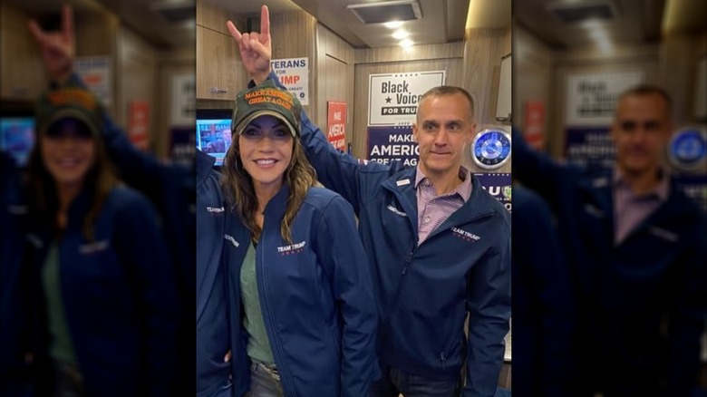 Corey Lewandowski and Kristi Noem wearing blue "Team Trump" jackets as Lewandowski makes the sign of the horns over Noem's head.