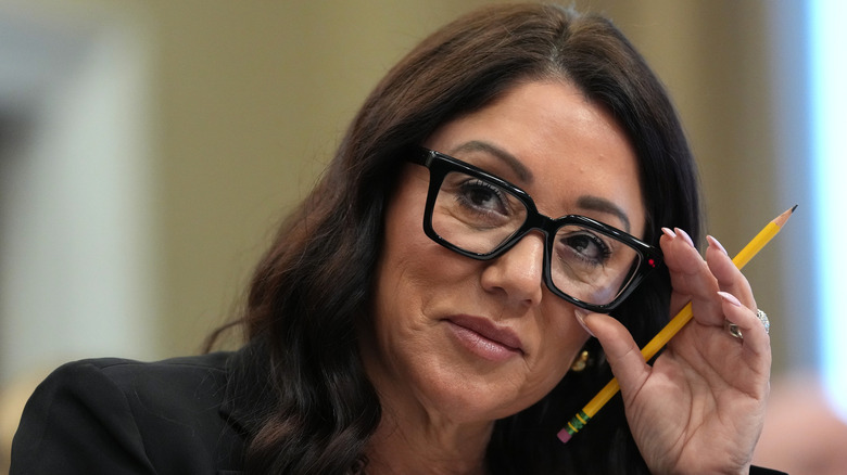 Lori Chavez-DeRemer smiles and adjusts her glasses while holding a pencil.