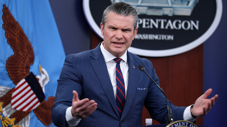 Pete Hegseth frowns and gestures with his palms up while wearing a blue suit and striped tie during a press conference.