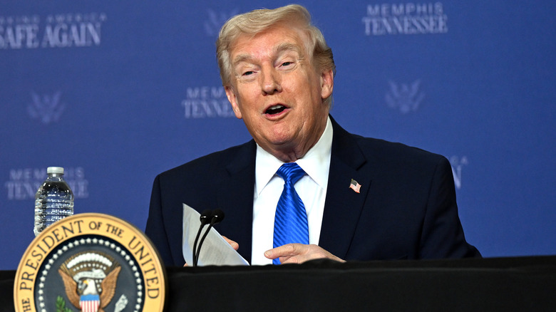 Donald Trump speaking at a podium in a black suit with a blue tie