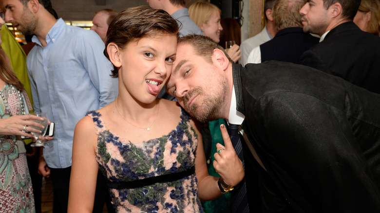 Young Millie Bobby Brown sticking her tongue out and pointing at David Harbour who rests his head on her shoulder.