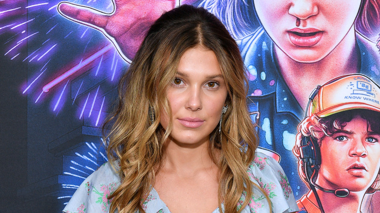Millie Bobby Brown at "Stranger Things" event standing in front of poster