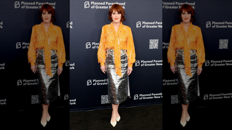 Molly Ringwald smiles in a orange top and metallic silver skirt