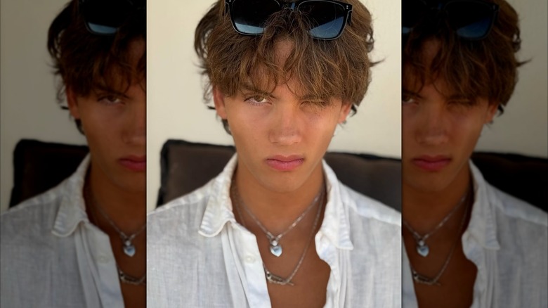 Closeup of Roman Stylianos Gianopoulos posing in white shirt, silver necklaces, and sunglasses atop his head