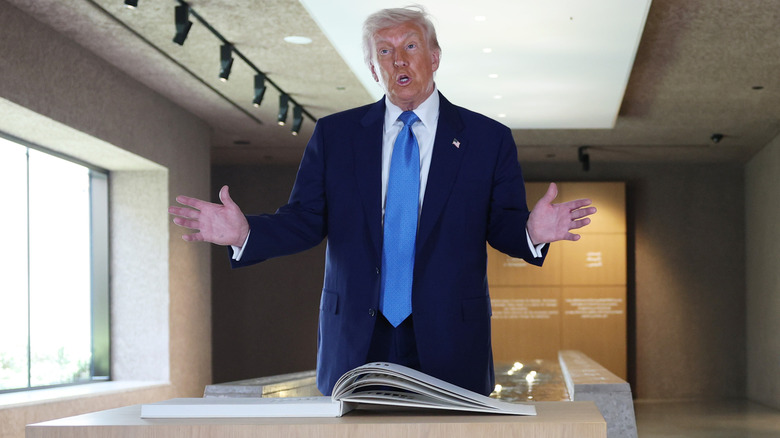 Donald Trump stands with his arms outstretched behind a table with a large book on it.