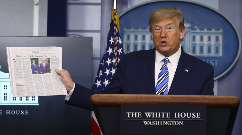 Donald Trump stands behind a lectern and holds a newspaper in the White House press briefing room.