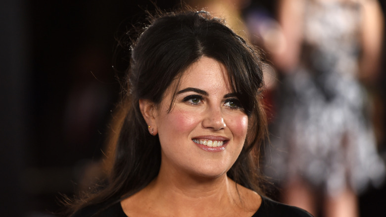 Monica Lewinsky with long hair and bangs, she wears a black dress with a blurred background.