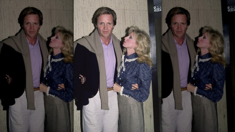 Jon Voight stares ahead at a camera while Morgan Fairchild looks up at him in a patterned blue blouse with lace trims and a wool gray skirt