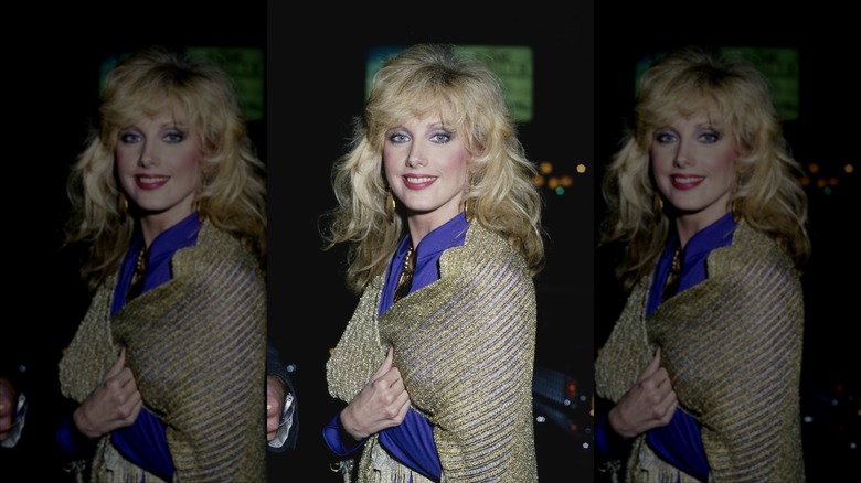 Morgan Fairchild holding a gold sparkly shawl over her blue silk blouse while smiling for the camera