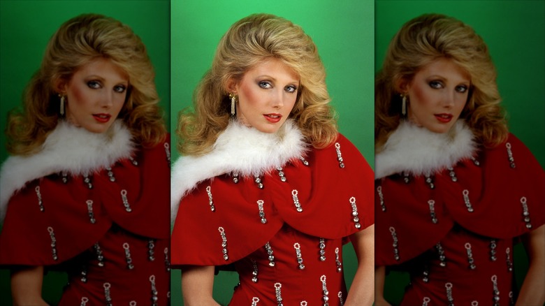 Morgan Fairchild posing against a green background in a red embellished shawl and dress with a fuzzy white trim along the neckline