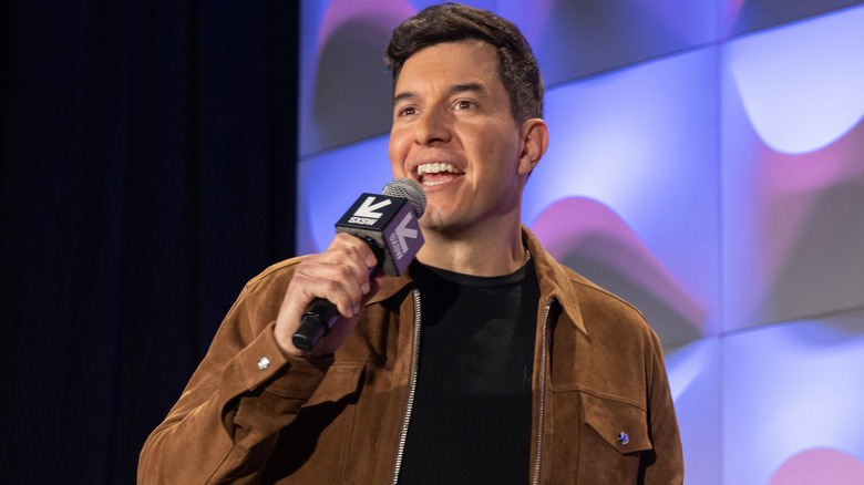 Tom Llamas speaking into a microphone in a brown suede jacket