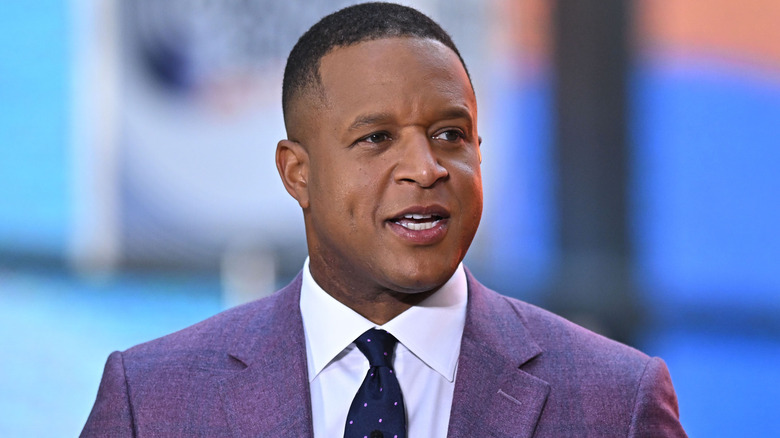 Craig Melvin speaking on TV in a grey suit with a white shirt and black polka dot tie