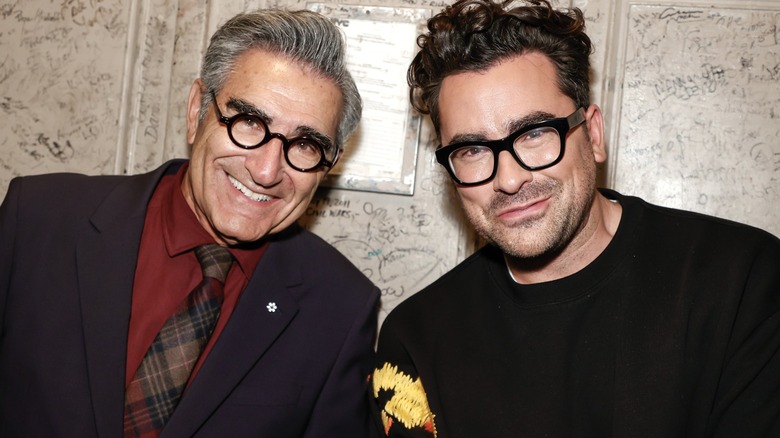 Eugene Levy and Dan Levy posing together in front of a wall of signatures