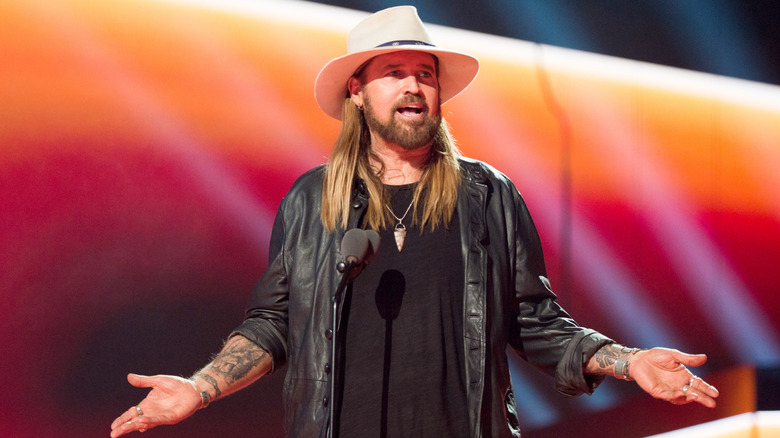Billy Ray Cyrus gesturing onstage in a black leather jacket and a wide-brimmed beige hat
