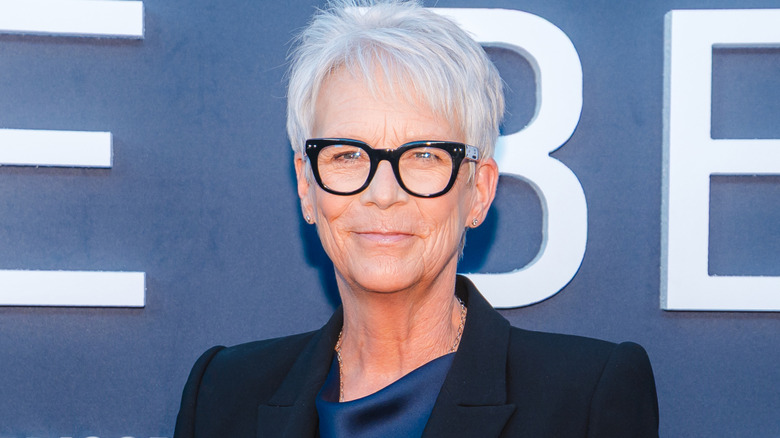 Jamie Lee Curtis smiling while posing on the red carpet in a black blazer and glasses