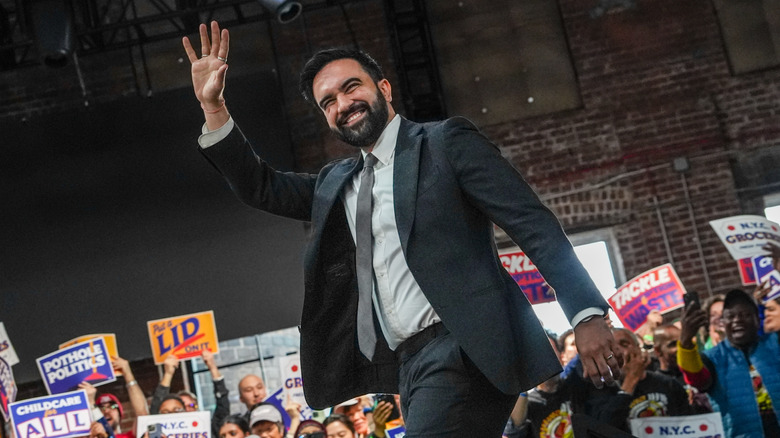 Zohran Mamdani waving and smiling at a big crow of supporters with signs