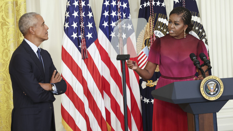 Michelle Obama delivering a speech while Barack Obama watches her with his arms crossed