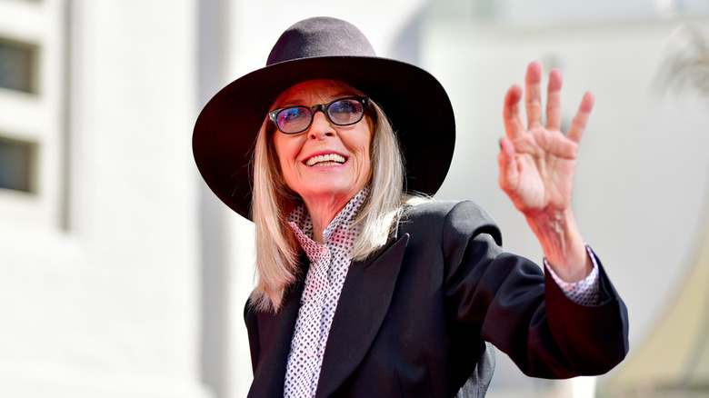 Diane Keaton waving in a black hat, glasses, and a black blazer