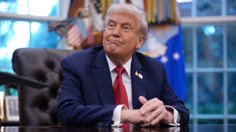 Donald Trump sitting in the Oval Office with his hands together looking smug