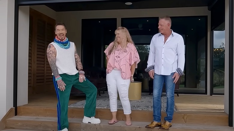 David Bromstad wearing green track pants and chunky beads, standing with two 'My Lottery Dream Home' guests