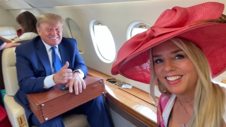Pam Bondi in wide-brimmed hat taking selfie on private plane with Donald Trump who's giving thumbs up and clutching briefcase