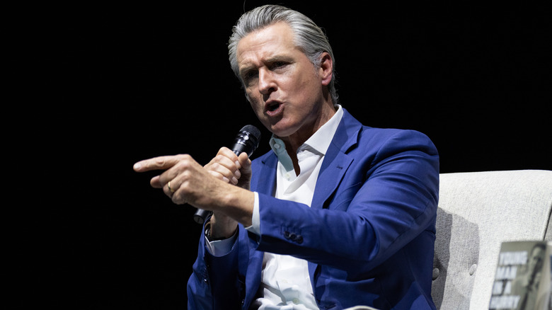 Gavin Newsom looking furious while speaking onstage in a blue blazer with an open-collared white shirt underneath