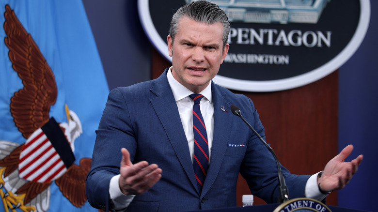 Pete Hegseth looking confused while speaking in front of a microphone in a navy suit