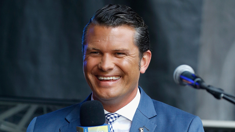Pete Hegseth smiling in a blue suit while holding a microphone.