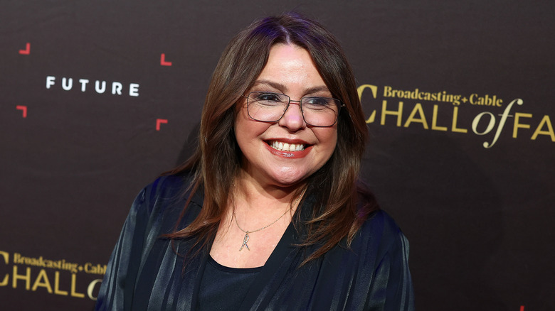 Rachael Ray in a navy outfit, in glasses, smiling and looking off to the right
