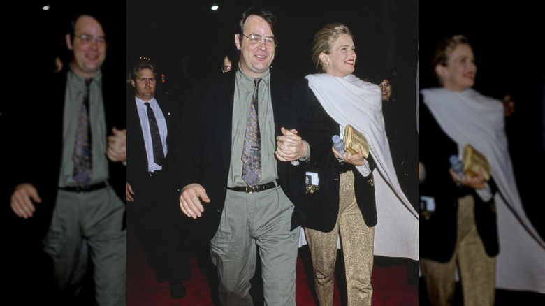 Dan Aykroyd, wearing a black blazer over a grey shirt with a patterned tie, holding hands with his wife, American actress Donna Dixon, who wears a white cape over a black blazer and gold trousers.