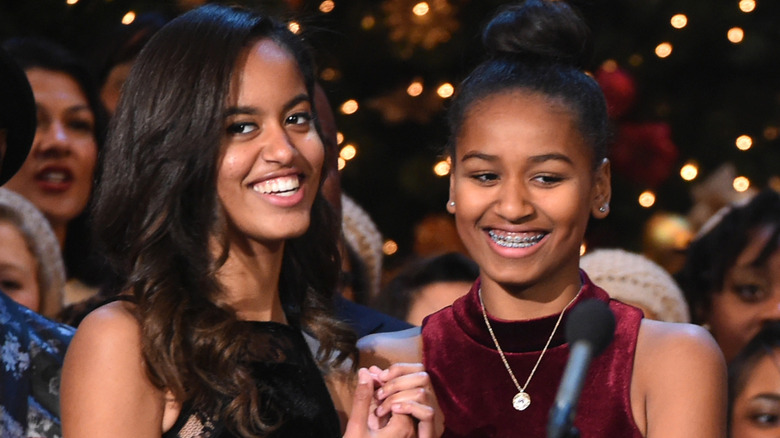 Malia Obama, her hair curled, in a black dress standing next to Sasha Obama in a red velvet dress with her hair in a bun.