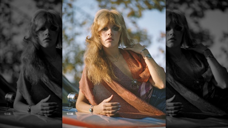 Stevie Nicks in 1975 posing on a Chevelle wearing an orange top and blue jeans and turquoise jewelery