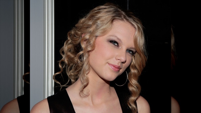 Taylor Swift with her head titled in a black dress with large hoop earrings.
