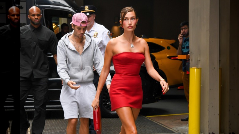 Justin Bieber in sweatshirt and shorts, walking alongside Hailey Bieber in red dress