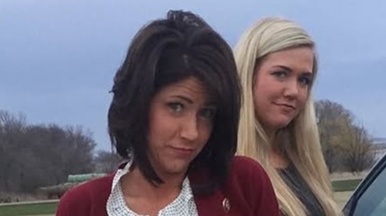 Kristi Noem smirking in a red sweater while in the middle of a parking lot.