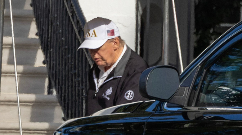 Donald Trump in a white hat, walking beside a black car.