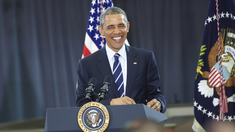 Barack Obama laughing while standing behind a podium in a navy suit