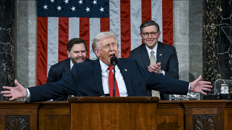 Donald Trump standing with his arms outstretched while addressing Congress with JD Vance and Mike Johnson applauding with wide grins behind him