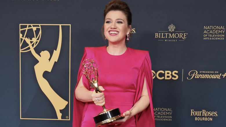 Kelly Clarkson in a pink dress with pronounced shoulder pads smiling for the cameras and posing with her Daytime Emmy Award